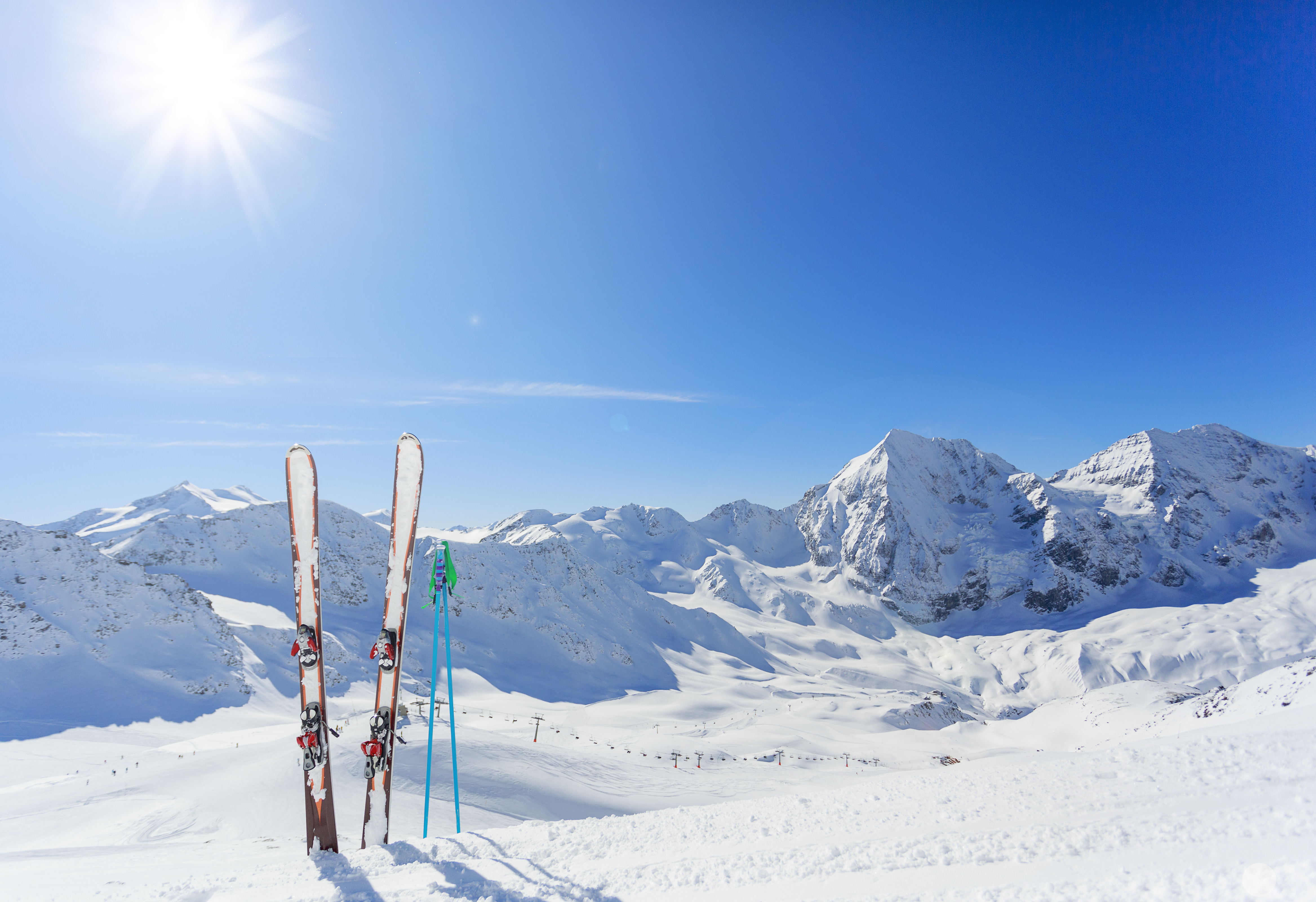 a pair of skis and ski poles dug into the snow with huge mountains and the bright sun behind