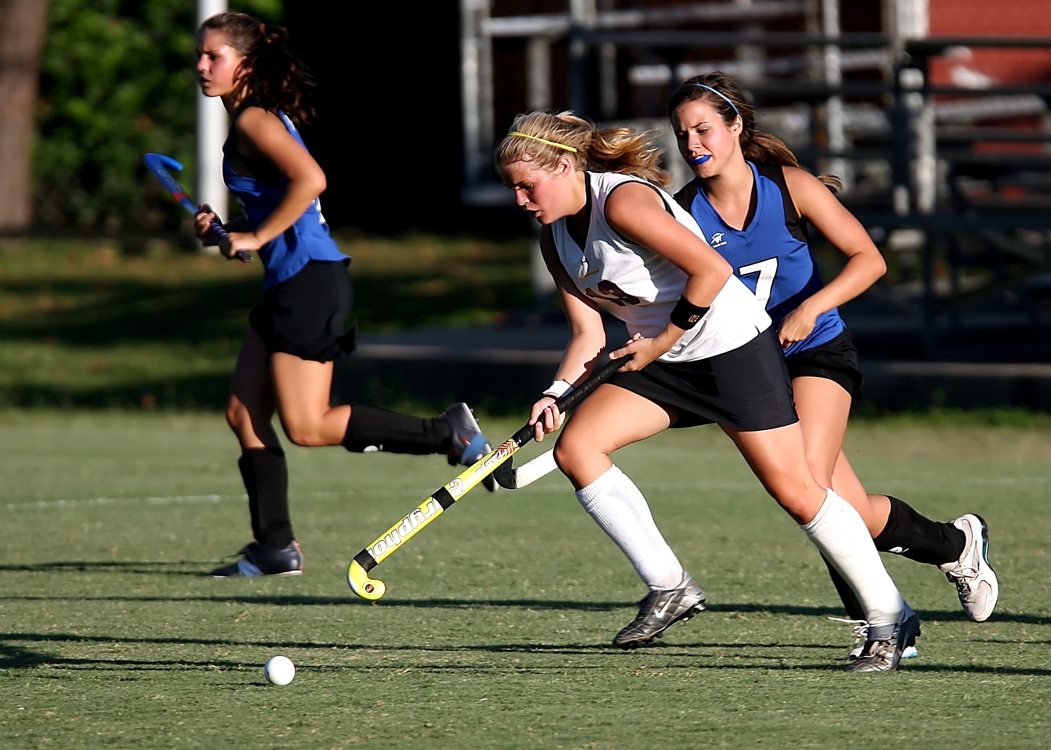 three players going after the ball in a field hockey game