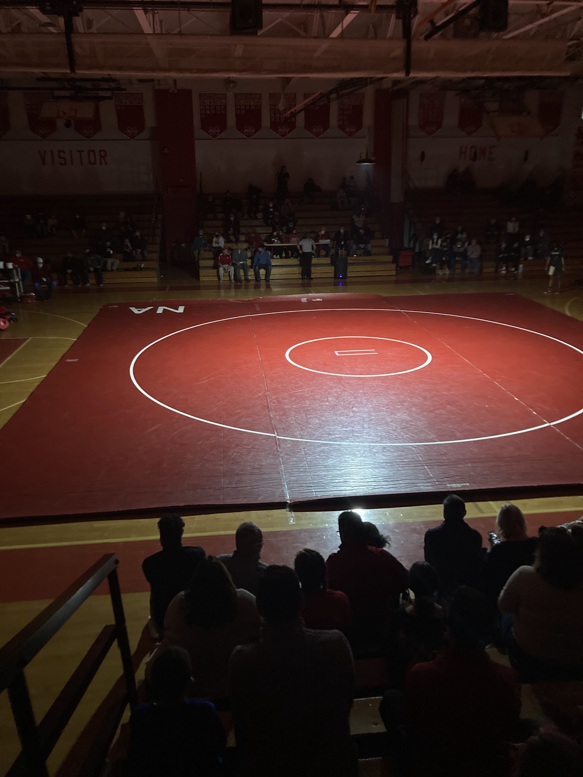 A wrestling mat is illuminated under a spotlight in a dimly lit gymnasium. Spectators are seated in the bleachers around the mat, with some sections of the bleachers marked as 'Visitor' and 'Home.' The red mat stands out against the darkened background, and a few people are standing near the edges of the mat.