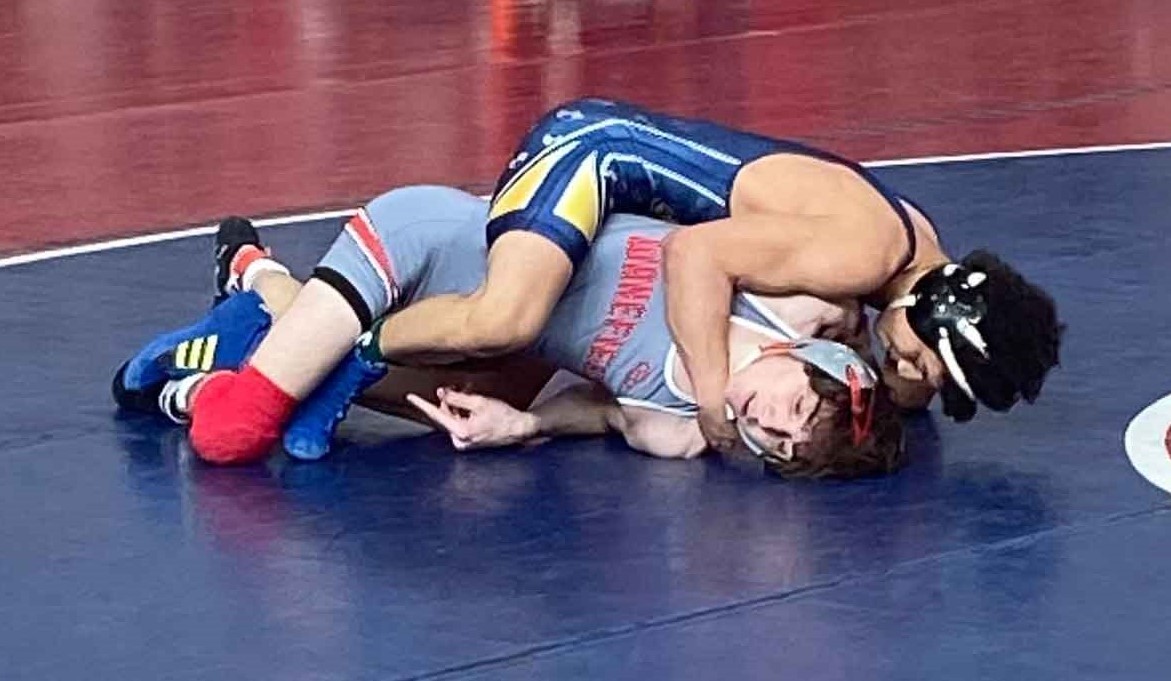 Two wrestlers are engaged in a match on a wrestling mat. The wrestler in blue and yellow is on top, holding down the wrestler in grey and red. The wrestler on the bottom has one arm pinned and is looking towards the camera with a grimace. The action is taking place on a blue mat with a red mat in the background.