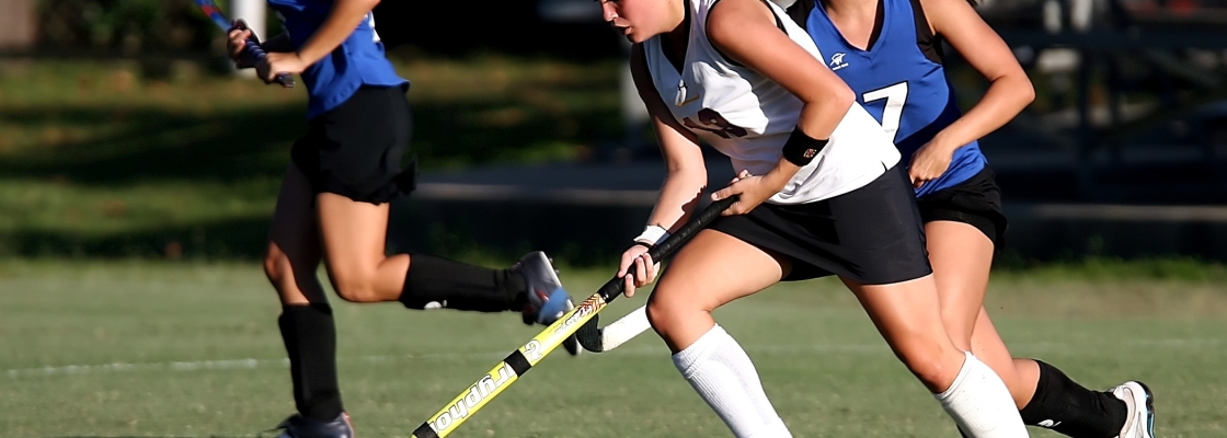 three players going after the ball in a field hockey game